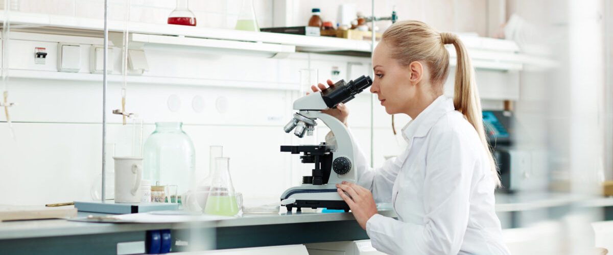 Researching Young woman in white-coat studying new virus in microscope